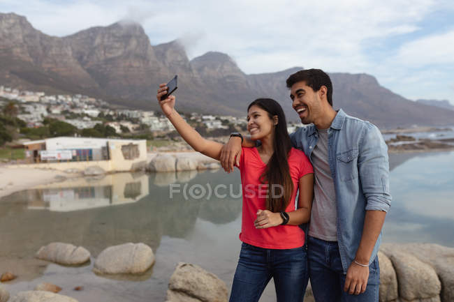 Vue de face rapprochée d'un jeune couple mixte souriant et prenant des selfies avec un smartphone, debout sur une plage avec la mer et les montagnes en arrière-plan — Photo de stock