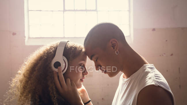 Vista lateral de uma jovem hispano-americana e uma jovem caucasiana usando fones de ouvido com os olhos fechados e as testas reunidas dentro de um armazém vazio — Fotografia de Stock