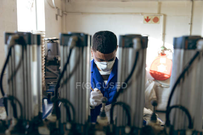 Front view of a young mixed race man wearing gloves and a face mask looking down and preparing a mixture at a factory making cricket balls, seen through equipment in the foreground. — Stock Photo