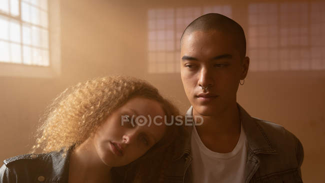 Front view of a young Caucasian female with curly hair looking intently at the camera while resting head on the shoulder of a young Hispanic-American man inside an empty warehouse — Stock Photo