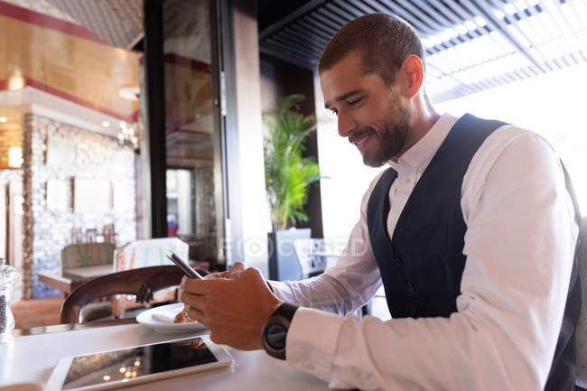 Vue de côté gros plan d'un jeune homme caucasien souriant utilisant son smartphone assis à une table à l'intérieur d'un café. Nomade numérique en mouvement . — Photo de stock