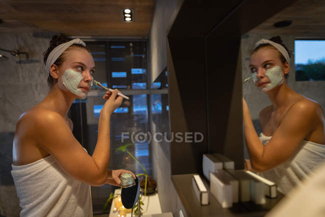 Вид сбоку молодой кавказки, смотрящей в зеркало и надевающей маску для лица кистью в современной ванной комнате . — стоковое фото