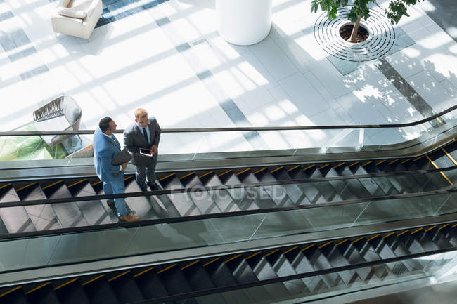 Visão de alto ângulo de empresários interagindo uns com os outros enquanto se deslocam para baixo na escada rolante no escritório . — Fotografia de Stock