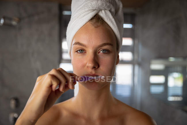 Портрет молодой белой женщины с завернутыми в полотенце волосами, чистящей зубы, смотрящей прямо в камеру в современной ванной комнате . — стоковое фото