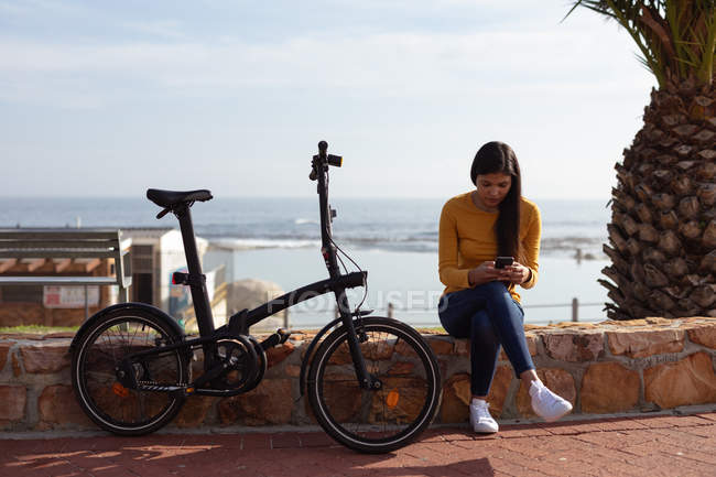 Vista frontal de una joven mestiza sentada afuera en una pared usando un teléfono inteligente con su bicicleta apoyada a su lado, una palmera y una vista al mar en el fondo - foto de stock