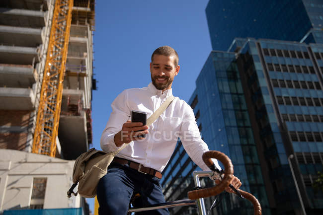 Vista frontal de cerca de un joven caucásico sonriente usando un teléfono inteligente, sentado en su bicicleta en una calle de la ciudad. Nómada digital en movimiento . - foto de stock