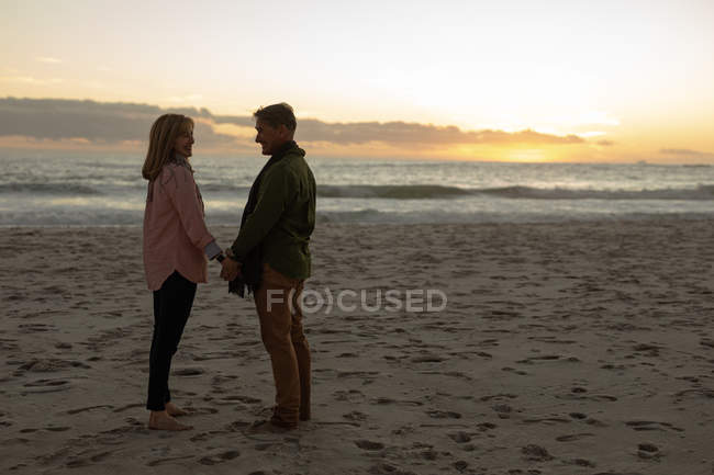 Вид збоку зрілих кавказьких чоловік і жінка тримає руки і стояти посміхаючись один на одного на пляжі біля моря на заході сонця — стокове фото