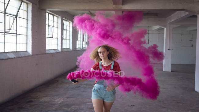 Frontansicht einer jungen kaukasischen Frau mit lockigem Haar, die in einer leeren Lagerhalle eine Rauchmaschine hält, die einen rosafarbenen Rauch produziert — Stockfoto