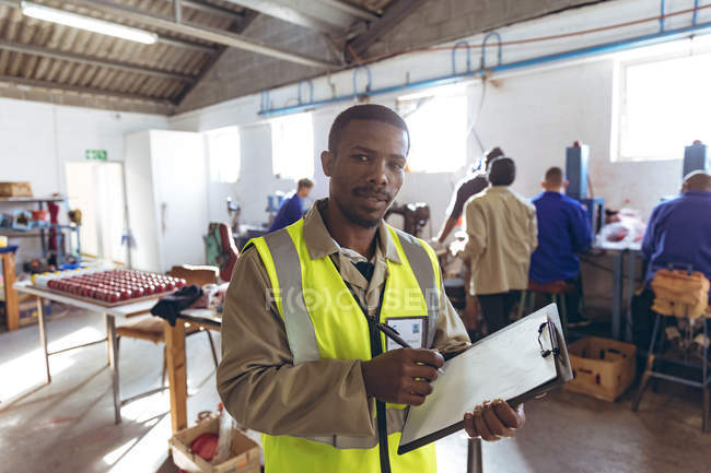 Retrato de perto de um jovem empresário afro-americano escrevendo em uma prancheta em uma fábrica de bolas de críquete, olhando para a câmera e sorrindo, com colegas de trabalho trabalhando em segundo plano . — Fotografia de Stock