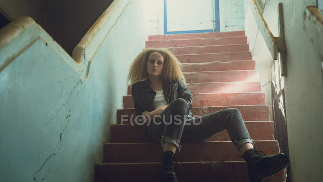 Низький кут зору молода кавказька жінка з Кучеряве волосся носіння шкіряна куртка сидить на сходах, дивлячись уважно на камеру — стокове фото