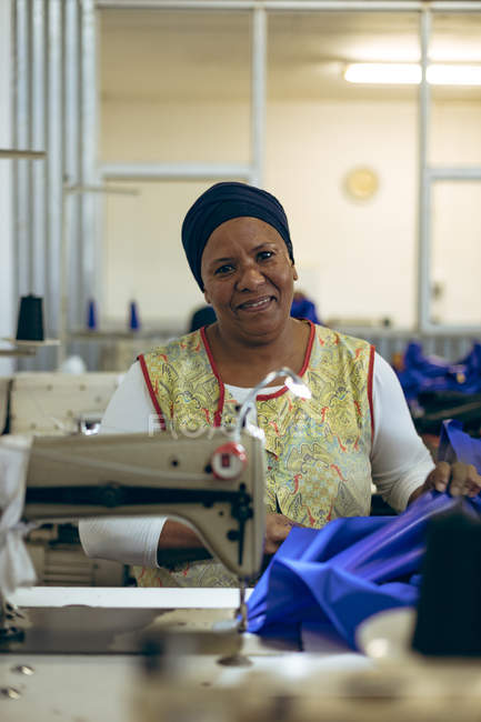 Portrait rapproché d'une femme métissée d'âge moyen utilisant une machine à coudre dans une usine de vêtements de sport, regardant vers la caméra et souriant . — Photo de stock