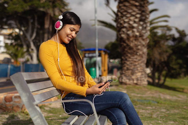 Вид збоку крупним планом усміхнена молода змішана жінка, що сидить на лавці в навушниках і використовує свій смартфон у парку, з пальмою на задньому плані — стокове фото