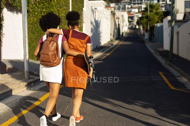 Vista trasera de dos hermanas adultas jóvenes de raza mixta con los brazos alrededor abrazándose mientras caminan en una calle bajo el sol, una con una mochila y la otra con un monopatín - foto de stock