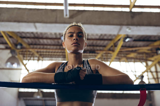 Низкий угол обзора женского боксера, опирающегося на веревки и смотрящего на камеру в боксерском ринге. Сильная женщина-боец в боксёрском зале тяжело тренируется . — стоковое фото