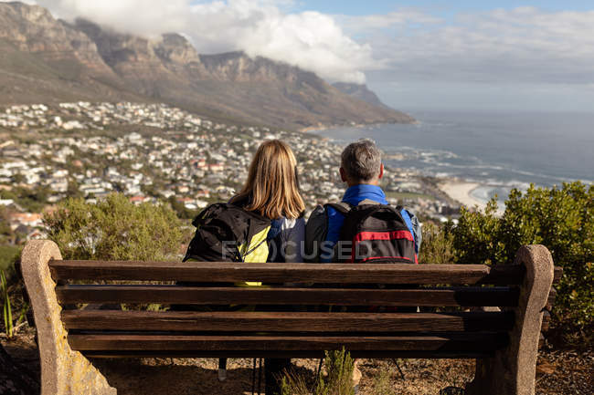 Vista trasera de una mujer caucásica madura y un hombre con mochilas sentadas en un banco y disfrutando de la vista durante una caminata . - foto de stock
