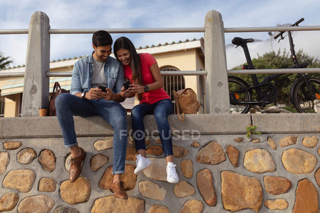 Vista frontal de una joven y sonriente pareja de raza mixta sentada afuera en una pared al sol mirando a sus teléfonos inteligentes - foto de stock