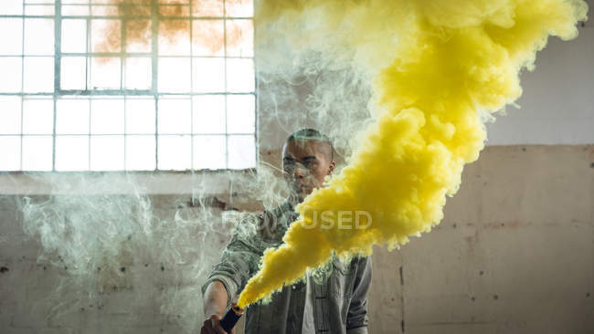 Vista frontal de un joven hispano-americano con una chaqueta gris sobre una camisa blanca mirando hacia otro lado de la cámara mientras sostiene una máquina de humo que produce humo amarillo dentro de un almacén vacío - foto de stock