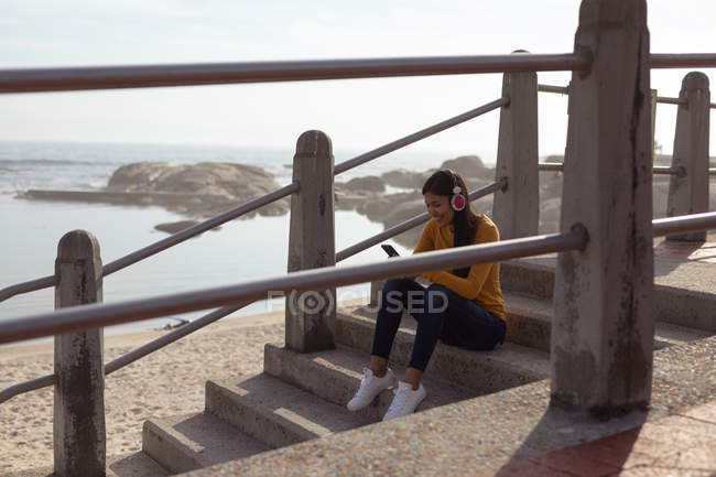 Vista lateral de una joven mujer de raza mixta sonriente sentada en escaleras con auriculares y usando su teléfono inteligente, con la playa y el mar en el fondo - foto de stock