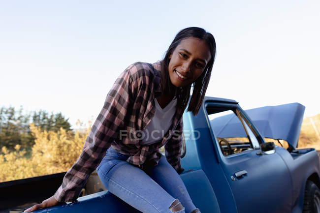 Vista frontal de cerca de una joven mujer mestiza sentada en la parte trasera de una camioneta sonriendo a la cámara durante una parada en un viaje por carretera
. - foto de stock
