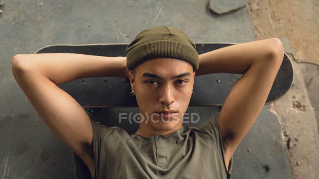 High angle view of a young Hispanic-American man with piercings wearing a dark grey shirt and beanie lying on the floor with head on a skateboard looking intently at the camera — Stock Photo