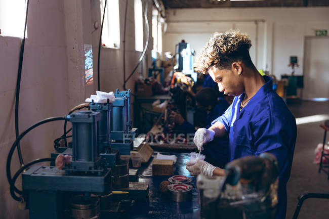 Side view of a young mixed race man wearing gloves and overalls preparing a mixture for the production line at a cricket ball factory, with colleagues operating machines in the background. — Stock Photo