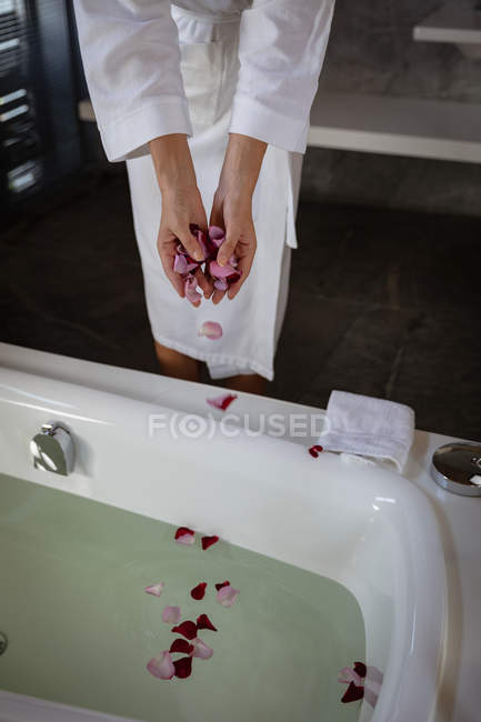 Metà sezione di donna indossa un accappatoio spargimento petali di rosa in un bagno pieno d'acqua in un bagno moderno
. — Foto stock