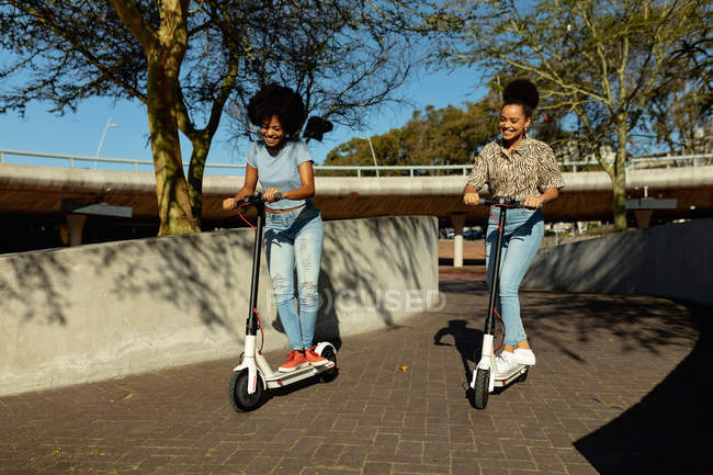 Vista frontal de dos hermanas adultas jóvenes de raza mixta montadas en scooters eléctricos en un parque urbano sonriendo - foto de stock
