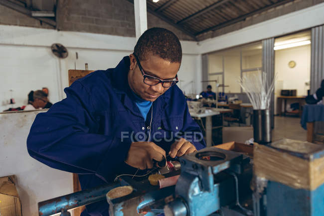 Vue de face gros plan d'un jeune Afro-Américain portant des lunettes debout sur un établi tenant du fil dans sa bouche et ses mains, cousant une balle dans un atelier d'une usine fabriquant des balles de cricket . — Photo de stock