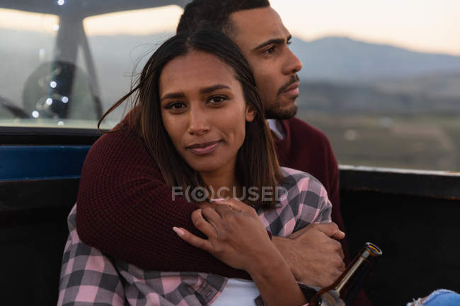Close up front view of a young mixed race couple sitting outside in the back of their pick-up truck, embracing and enjoying the view at sundown during a stop off on a road trip. — Stock Photo