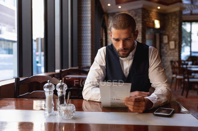 Vista frontal de perto de um jovem caucasiano sentado em uma mesa em um café olhando para o menu com seu smartphone na mesa ao lado dele. Digital Nomad em movimento . — Fotografia de Stock