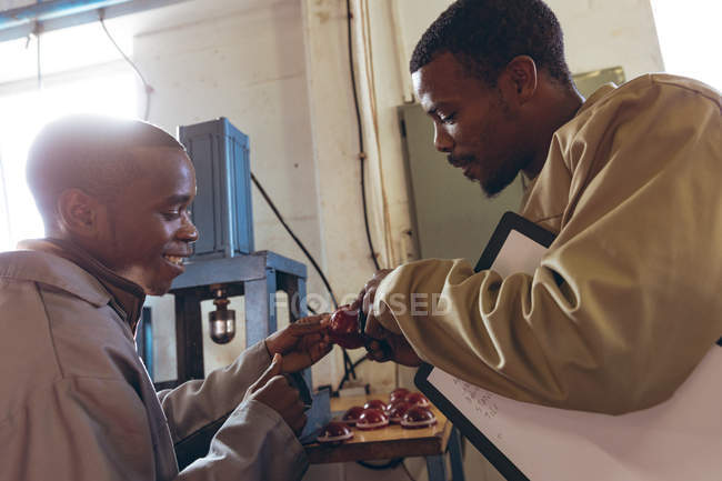 Vista lateral de perto de um jovem gerente masculino afro-americano segurando uma prancheta conversando com um jovem afro-americano trabalhando em uma fábrica de bolas de críquete e inspecionando seu trabalho . — Fotografia de Stock