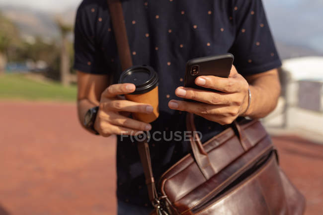 Vista frontal sección media del hombre con un bolso de hombro, sosteniendo un café para llevar y utilizando un teléfono inteligente fuera en el sol - foto de stock