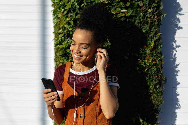 Vista frontal de cerca de una joven mujer de raza mixta sonriente usando su teléfono inteligente con auriculares, parada al aire libre bajo el sol - foto de stock