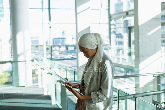Vue latérale de femme d'affaires en hijab travaillant sur tablette numérique près de rampe dans un bureau moderne . — Photo de stock