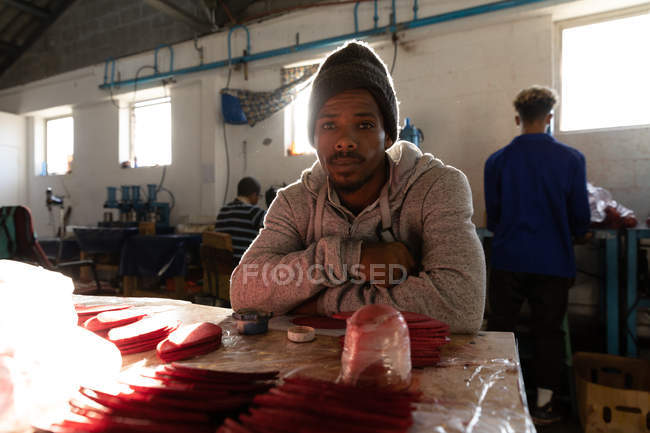 Portrait close up of a young African American man sitting at a workbench in a factory making cricket balls, looking to camera and smiling. — Stock Photo