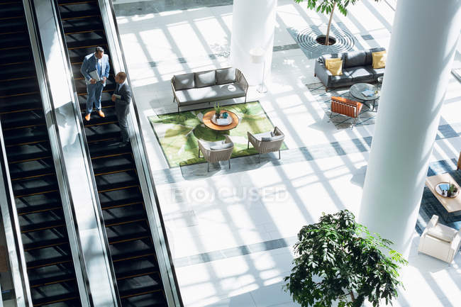 Hochwinkelaufnahme von Geschäftsleuten, die miteinander interagieren, während sie sich auf der Rolltreppe im Büro nach unten bewegen. — Stockfoto