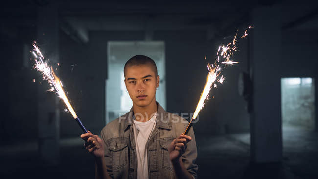 Front view of a young Hispanic-American man wearing a grey jacket over a white shirt looking intently at the camera while holding lighted sparkles on both hands inside an empty warehouse — Stock Photo