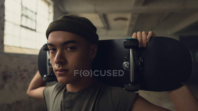 Close up view of a young Hispanic-American man with piercings wearing a dark grey shirt and beanie while holding a skateboard over shoulder and looking intently at the camera while standing inside an empty warehouse — Stock Photo