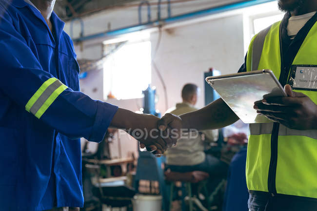 Side view mid section of male colleagues shaking hands at a cricket ball factory, one is holding a tablet computer, with people working at machines in the background. — Stock Photo