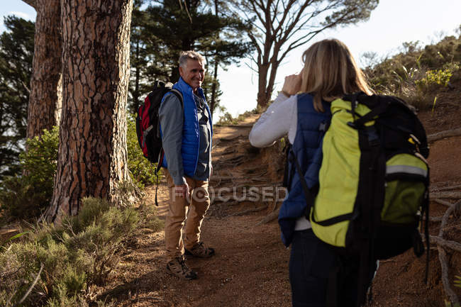 Rear view of a mature Caucasian woman and side view of mature Caucasian man both wearing backpacks walking single file along a trail during a hike, the man has turned and is smiling at the woman — Stock Photo