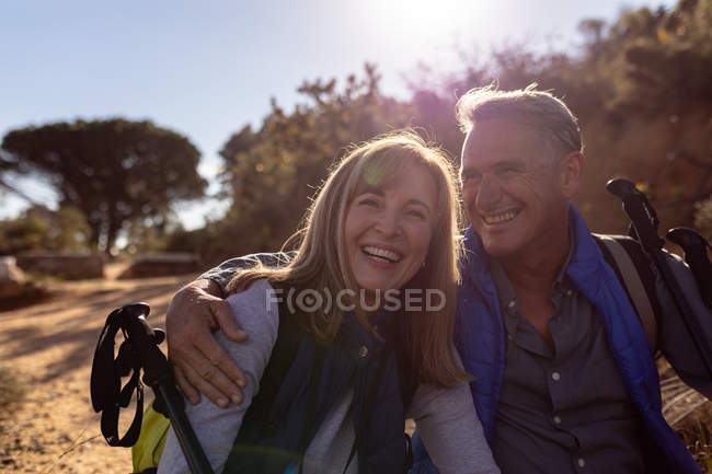 Вид спереди на взрослую белую женщину и мужчину в рюкзаках и с палками для нордической ходьбы, смеющиеся и обнимающиеся во время похода, освещенные солнечным светом — стоковое фото