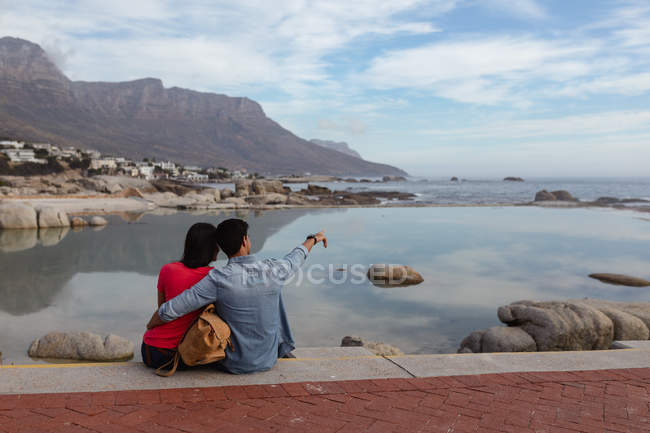 Vista trasera de una joven pareja de raza mixta sentada afuera en una pared abrazando y mirando hacia el mar, el hombre señala a la distancia - foto de stock