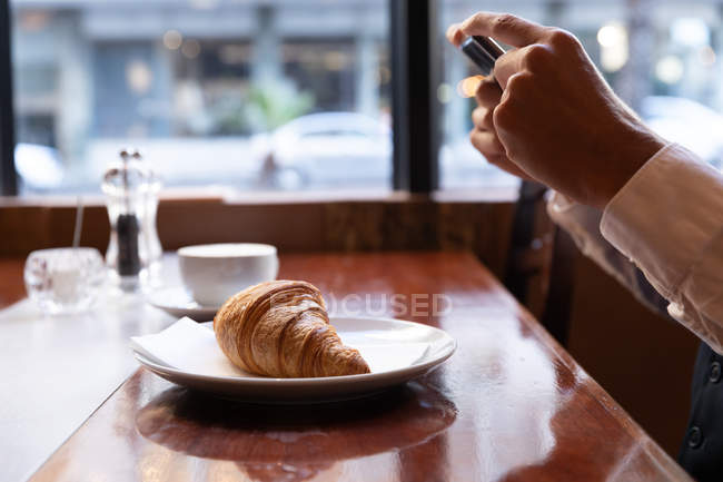 Вид сбоку закрывает руки человека, фотографирующего свой круассан со смартфоном, сидящего за столом в кафе. Цифровая реклама на ходу . — стоковое фото