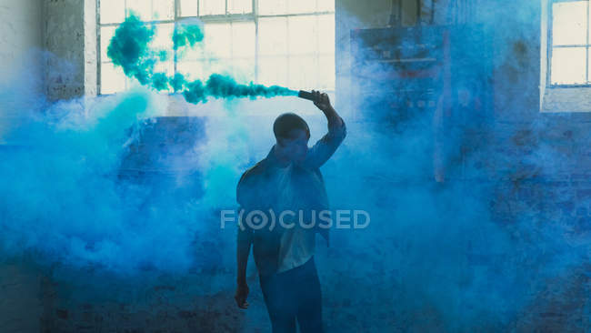 Vista frontal de um jovem hispânico-americano vestindo uma jaqueta cinza sobre uma camisa branca segurando um fabricante de fumaça produzindo fumaça azul dentro de um armazém vazio — Fotografia de Stock