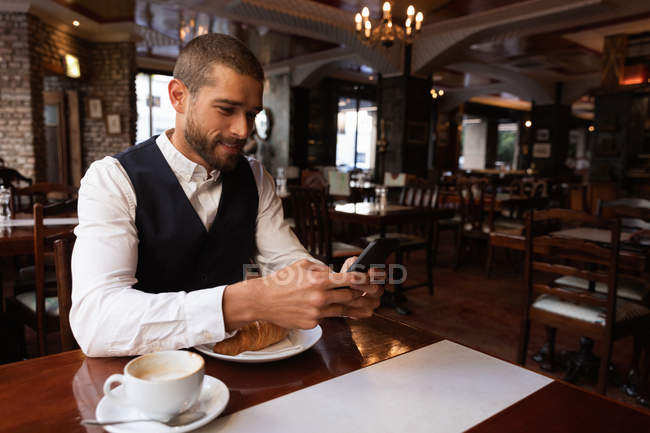 Vista frontal de cerca de un joven caucásico usando su teléfono inteligente sentado en una mesa con café y un croissant dentro de un café. Nómada digital en movimiento . - foto de stock