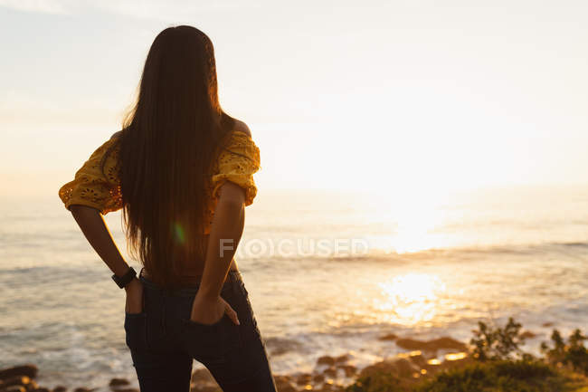 Вид ззаду крупним планом молода змішана жінка з довгим волоссям, що стоїть на пляжі з її руками в задніх кишенях її джинсів, дивлячись на море на заході сонця — стокове фото