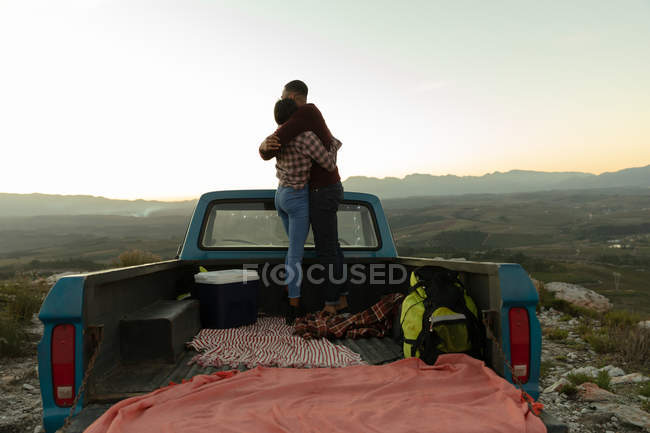 Vue arrière d'un jeune couple de course mixte debout à l'arrière de sa camionnette, embrassant et appréciant la vue au coucher du soleil lors d'un arrêt sur la route . — Photo de stock