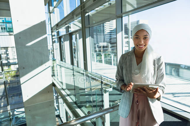 Glückliche Geschäftsfrau im Hijab blickt in die Kamera, während sie an einem digitalen Tablet in der Nähe des Geländers im Büro arbeitet. — Stockfoto