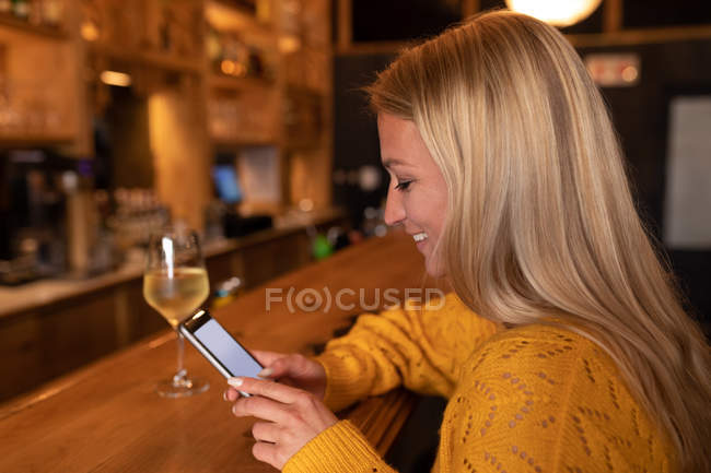 Vista lateral de una joven mujer caucásica feliz relajarse de vacaciones en un bar, beber vino y usar un teléfono inteligente - foto de stock