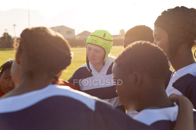 Sobre la vista del hombro de un equipo de jóvenes jugadoras de rugby multiétnicas adultas de pie en un campo de rugby con los brazos conectados preparándose para un partido de rugby - foto de stock